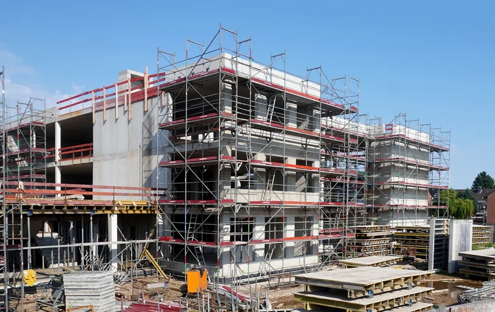 Seitlicher Blick von der Bodelschwinghstraße auf den Rohbau der neuen Petrus-Canisius-Grundschule in Weeze im Juni 2026. Mehrere Gebäudeteile sind bereits bis in die oberen Etagen gewachsen und von Gerüsten umgeben. Im Vordergrund liegen Schalungselemente und Baumaterialien, während die künftige Form des Schulgebäudes zunehmend erkennbar wird.